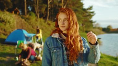 Güneş ışığında kızıl saçlı çekici genç bir kadın kameraya bakarken mutlu olur. Arka planda arkadaşları piknik malzemelerini yerleştiriyor. Ormanda gölün yanında dur. Kapatın. Yavaş çekim