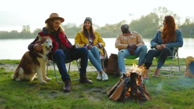 Ağır çekim arkadaşlar doğada kamp ateşinin yanında oturuyorlar. Gençler oyunlar hakkında konuşurlar, köpeklerin gülümsemesiyle mutlu olurlar. Tatil turizmi macerası. Kapat.