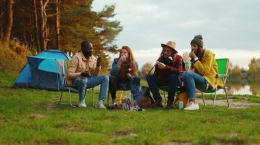 Mutlu arkadaşlar kamp ateşi etrafında oturur ve renkli şekerlemeler kızartırlar. Ormandaki gençler. Kamp yapmak. Piknik. Yavaş çekim