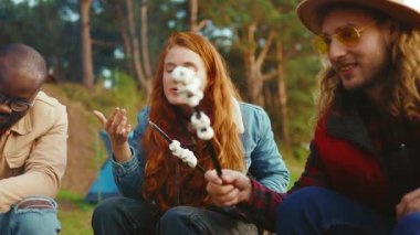 Genç arkadaşlar kamp ateşinin etrafında oturup renkli marşmelovları kızartıyorlar. Ormandaki gençler. Kamp yapmak. Piknik. Yavaş çekim