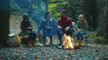 Gülümseyen aile sonbahar akşamı kamp ateşinin yanında oturur. Ebeveynleriyle birlikte çocuklar ormanda dinleniyorlar. Çocukluk ilişkisi kamp kuruyor. Kapatın. Yavaş çekim