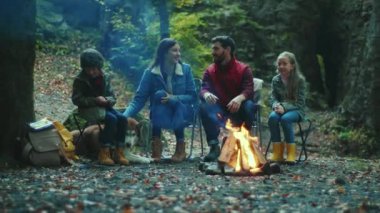 Komik aile sonbahar akşamı kamp ateşinin yanında oturur. Ebeveynleriyle birlikte çocuklar ormanda dinleniyorlar. Çocukluk ilişkisi kamp kuruyor. Kapatın. Yavaş çekim