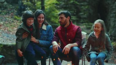 Gülümseyen aile sonbaharda kamp ateşinin yanında oturur. Ebeveynleriyle birlikte çocuklar ormanda dinleniyorlar. Çocukluk ilişkisi kamp kuruyor. Kapatın. Yavaş çekim