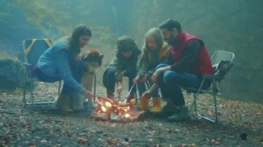 Köpekli mutlu bir aile, sonbahar akşamı ormanda kamp ateşinin etrafında oturur. Ormandaki bir yangında sosis kızartmak. Arkadaşlık şömineyi pişirir. Kapatın. Yavaş çekim
