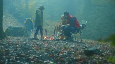 Köpekli güzel bir aile, sonbahar akşamı ormanda kamp ateşinin etrafında oturur. Ormandaki bir yangında sosis kızartmak. Arkadaşlık şömineyi pişirir. Kapatın. Yavaş çekim