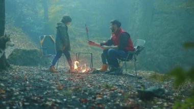 Köpekle gülen aile, sonbahar akşamı ormanda kamp ateşinin etrafında oturur. Ormandaki bir yangında sosis kızartmak. Arkadaşlık şömineyi pişirir. Kapatın. Yavaş çekim