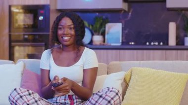 Siyah oturma odasındaki kanepede oturan Afrikalı Amerikalı kadın. Geceleri televizyon izlerken gülümseyen uzaktan kumandayla kanal değiştirmek. Yavaş çekim