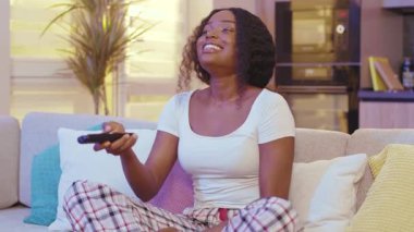 Geceleri, karanlık oturma odasındaki kanepede oturan Afrikalı Amerikalı kadın. Televizyon izlerken gülümseyen uzaktan kumandayla kanal değiştirmek. Yavaş çekim
