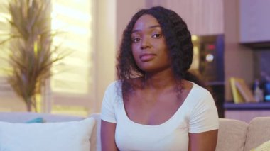Şirin, güzel, mutlu, Afro-Amerikan genç kadın evinde, kanepede oturan kameraya bakıyor. Karantina zamanı. Evde kal. Kapatın. Yavaş çekim