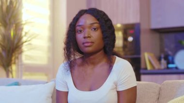 Ciddi, güzel, Afro-Amerikan genç bir kadın kameraya bakıyor. Kollarını sallıyor ve evinde, kanepede oturuyor. Karantina zamanı. Evde kal. Kapatın. Yavaş çekim
