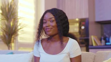 Güzel bir afro-amerikan genç bayanı portresi. Kameraya bak. Evde, kanepede oturuyor. Karantina zamanı. Evde kal. Kapatın. Yavaş çekim