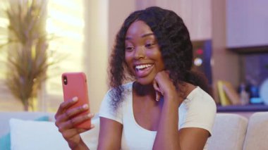 Mutlu Afrikalı Amerikalı genç kadın rahat rahat koltuğa otur ve gece evde telefonunu kullan. Rahat bir sosyal medya. Kapatın. Yavaş çekim
