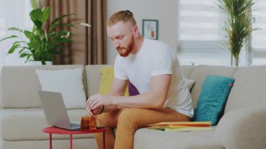 Genç yakışıklı adamın portresi kanepede otururken dizüstü bilgisayar kullanıyor. Çay iç. Evde çalış. İnternet teknolojisi. Karantina zamanı. Kapatın. Yavaş çekim