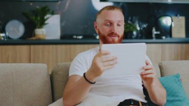 Tablet bilgisayar kullanan yakışıklı bir adam. Görüntülü konuşma. Evdeki kanepede oturmak. Çevrimiçi teknoloji aygıtı iletişim karantinası.