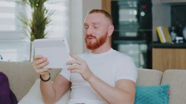 Tablet bilgisayar kullanan yakışıklı bir adam. Görüntülü konuşma. Evdeki kanepede oturmak. Çevrimiçi teknoloji aygıtı iletişim karantinası.