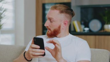 Evdeki kanepede oturan yakışıklı adam portresi. Akıllı telefon kullanarak dokunarak ve araklayarak. Sosyal medya interneti. Etrafına bak. Bağlantı iletişimi. Yavaş çekim