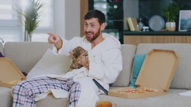 Kedisi olan sevimli bir genç adam TV seyrediyor ve pizza yiyor. Kanepede oturup gülümsemek. Dinlenme sinema kanalları dinleniyor. Portre. Kapatın. Yavaş çekim.