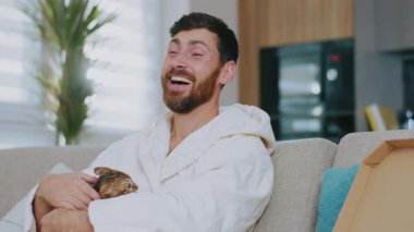 Pizza yiyen kedili genç bir adam portresi. Kanepede oturup gülümsemek. Dinlenme sinema kanalları dinleniyor. Portre. Kapatın. Yavaş çekim.