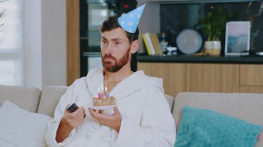 Üzgün genç adam doğum gününü tek başına kutluyor. Pasta izleyip televizyon izliyor. Karantina sırasında oturma odasındaki kanepede oturmak. Evde kalıp Coronavirus 'u altüst etmek. Yavaş çekim