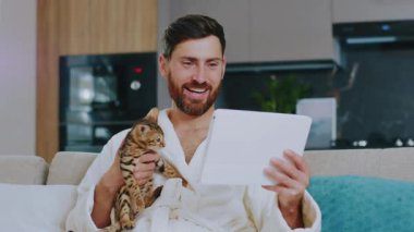 Evde kedisi olan mutlu yakışıklı bir adam arkadaşıyla görüntülü konuşma yapıyor. Evde kal. İletişim konferansı sosyal medya portresi. Yavaş çekim