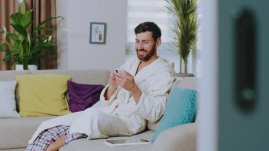 Ev kıyafetleri içindeki yakışıklı genç adam koltukta oturmuş gülümsüyor ve telefonu kullanıyor. Mutlu ol. Evde kal. Sosyal medya. İletişim konferansı portresi. Yavaş çekim