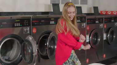 Genç ve komik sarışın kadın dansı çamaşırhanedeki çamaşır sepetinden çamaşır makinesine koyuyor. Ev hanımı ev işi, ev işi, ev içi çamaşır yıkama rutini. Yavaş çekim