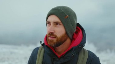 Yeşil örgü şapkalı, gülümseyen genç sakallı adam kar ormanında mutlu bir şekilde kameraya bakıyor. Çekici bir doğa. Kapatın. Yavaş çekim