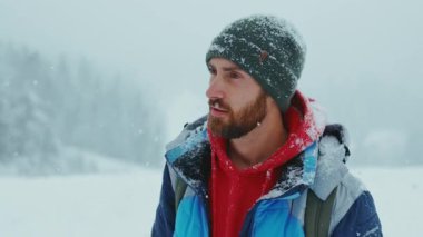 Yeşil örgü şapkalı yakışıklı genç sakallı adamın portresi kar ormanlarında gülümseyen kameraya bakıyor. Çekici bir doğa. Kapatın. Yavaş çekim