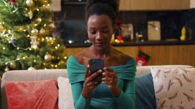 Yakın plan Afro-Amerikan güzel genç bayan kanepede oturuyor ve akıllı telefonda daktilo kullanıyor. Mutlu ol. Yeni yıl gecesi. Tatil kutlaması. Noel ağacı. Hediye. Yavaş çekim