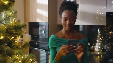 Mutlu Afrikalı Amerikalı genç bir kadının portresi Noel ağacının yanında duruyor ve cep telefonuna mesajlar yazıyor. Noel zamanı. Kış büyülü atmosferi kutluyor. Yavaş çekim