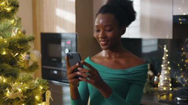 Gülümseyen Afrikalı Amerikalı genç kadın Noel ağacının yanında duruyor ve cep telefonuna mesajlar yazıyor. Noel zamanı. Kış büyülü atmosferi kutluyor. Yavaş çekim
