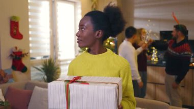 Afro-Amerikan güzel, mutlu, genç bir kadın. Gülümsemesine bakın. Noel hediye kutusunu dekore edilmiş odada tutun. Yeni yıl. Tatil kutlaması. Noel ağacı. Yavaş çekim