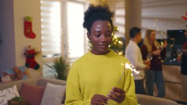 Noel ağacında süslü bir odada elinde maytapla gülümseyen geleneksel bayrama bakan Afrikalı Amerikalı genç bir kadın. Kutlama. Mutlu noeller. Kış zamanı. Yavaş çekim