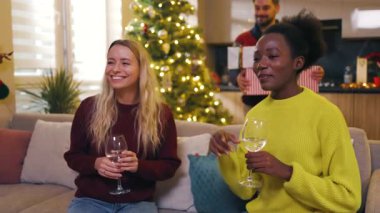 Çok ırklı iki kadın arkadaş noel partisi yapıyorlar ellerinde şarap tutuyorlar ev dansında kanepede oturuyorlar. Geleneksel tatil. Kutlama. Mutlu noeller. Kış zamanı. Yavaş çekim