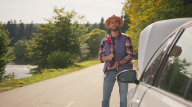 Üzgün gezgin hippi adam kaputu açık kırık bir arabanın yanında duruyor. Arızalı stresli sürücü boş yolda acil onarım için bekliyor..