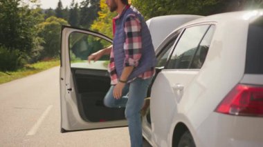 Hüsrana uğramış genç sürücü hippi sakallı beyaz adam arabayı durduruyor, yol kenarında duruyor ve motor kaputunu kontrol ediyor. Yol kazası. Sorun var..