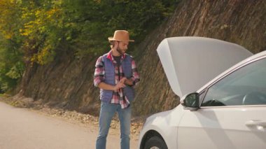 Üzgün hippi, sakallı, beyaz, sinirli adam arabanın kaputunu açıyor, boş kırsal yolda duruyor ve yardım bekliyor. Acil durum. Stres yüzünden. Araba yarışı. Araba arızası.