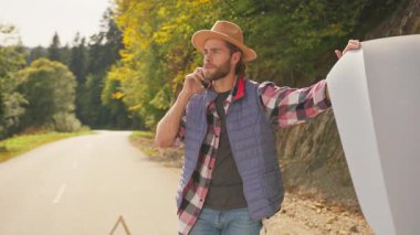 Sakallı genç turist arabanın kaputunu açıyor, acil servisi arıyor ve motor hasarı sorununu açıklıyor. Araba arızası. Araba durağı.