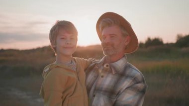 Aile fotoğrafı. Yakışıklı, beyaz, orta yaşlı, sakallı, çiftçi bir baba. Güzel, sevimli bir çocuğu elinde tutuyor, dışarıda gülümsüyor. Baba ve oğul. Gün batımı. Köy doğası.