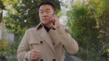 Hüsrana uğramış genç Asyalı işletme müdürü telefonda kızgın bir şekilde konuşuyor ve anlaşmazlık hakkında konuşuyor. Sokak manzaralı. Dışarı çıkın. Konuşma.
