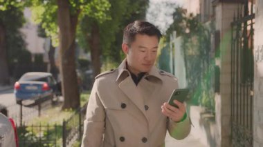 Asyalı çekici iş adamı mahallede akıllı telefon kullanıyor. Mutlu yakışıklı ve başarılı zengin adam paltolu gezgin yerler arıyor..