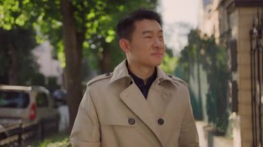 Çekici genç Çinli yetişkin bir adam güneşli ve güzel bir mahallede mutlu bir şekilde yürüyor. Başarılı bir insan. İş adamı. Asya etnik kökeni.