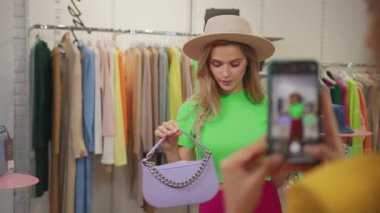 Birkaç kız arkadaş modern alışveriş merkezinde moda video bloğu kaydediyor. Sarışın, kendine güveni tam genç bir kadın mağazadaki şık kıyafetleri inceliyor..