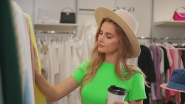 Genç, hoş görünümlü, beyaz saçlı, kahve fincanı tutan, alışveriş merkezindeki kıyafetlere bakan bir kadın. Alışverişkolik kavram.