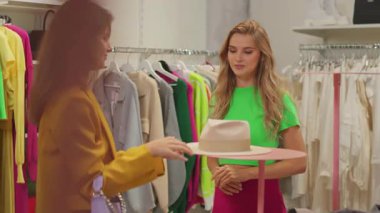 Pahalı dükkanda alışveriş yapan iki güzel kadın. Sarışın mutlu kız şık bir şapka takmaya çalışıyor. Arkadaşız. Alışveriş kolikleri.