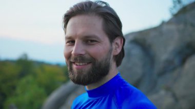 Yakından çekilen portre yakışıklı sakallı beyaz erkek sporcu mavi kıyafetli dışarıda kameraya neşeyle gülümsüyor. Sporcu suratı. Açık hava eğitimi.