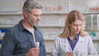 Kafkasyalı yetişkin bir adam eczaneye gidip antiviral ilaç ya da vitaminler hakkında kadın danışmanla konuşuyor. Modern eczacı. Eczane.