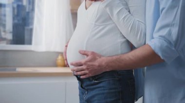 Genç koca elleriyle hamile karısına sarılıyor karnına dokunuyor ve birlikte eğleniyor. Mutfakta bir çift var. Aile hayatı. Sevgi dolu anlar.