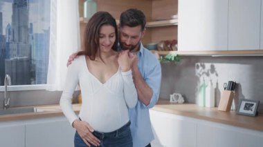 Sakallı, heyecanlı baba kocasından oluşan neşeli, genç, beyaz bir aile hamile karısına sarılıyor ve bebek bekliyor. İlişki hedefleri. Ebeveynlik.