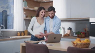 Mutfakta defter kullanan iki kişilik beyaz bir aile. Yakışıklı kocası olan hamile bir kadın çocuk kıyafetleri almak için internetten alışveriş yapıyor. Ebeveynlik.